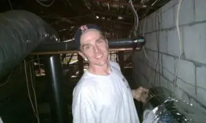 A man in white shirt and baseball cap standing next to pipe.